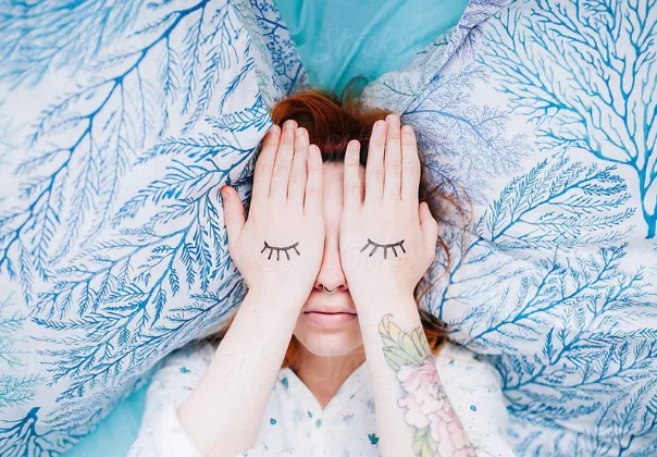 Women lying down on a bed with her hands covering her eyes and closed eyes drawn on her hands